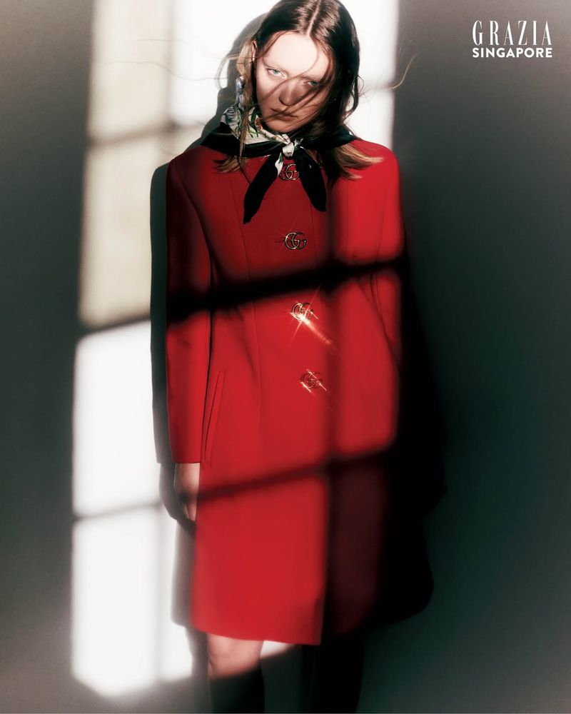 Model in a red coat with a dark headscarf standing against a wall with strong window light patterns for Grazia Singapore