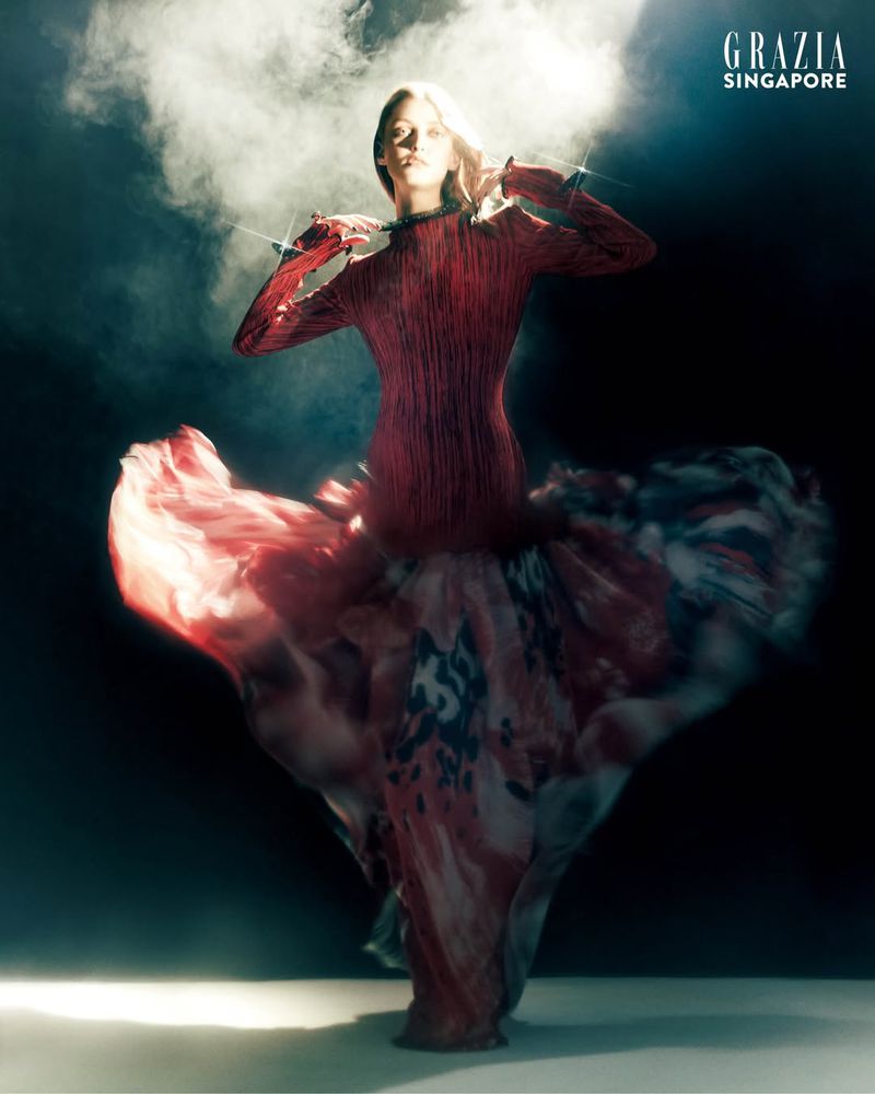 Model Polly Domashych in a red dress with a swirling patterned skirt against a dark smoky background with backlighting.