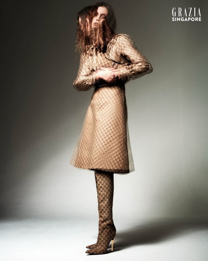 Fashion editorial shot of a woman in a patterned gold dress and matching boots in a photo studio with soft lighting and a grey background.
