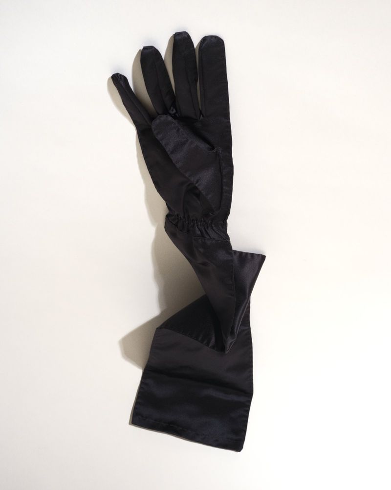 Long black satin evening glove displayed vertically against a pale neutral background, casting a soft shadow.