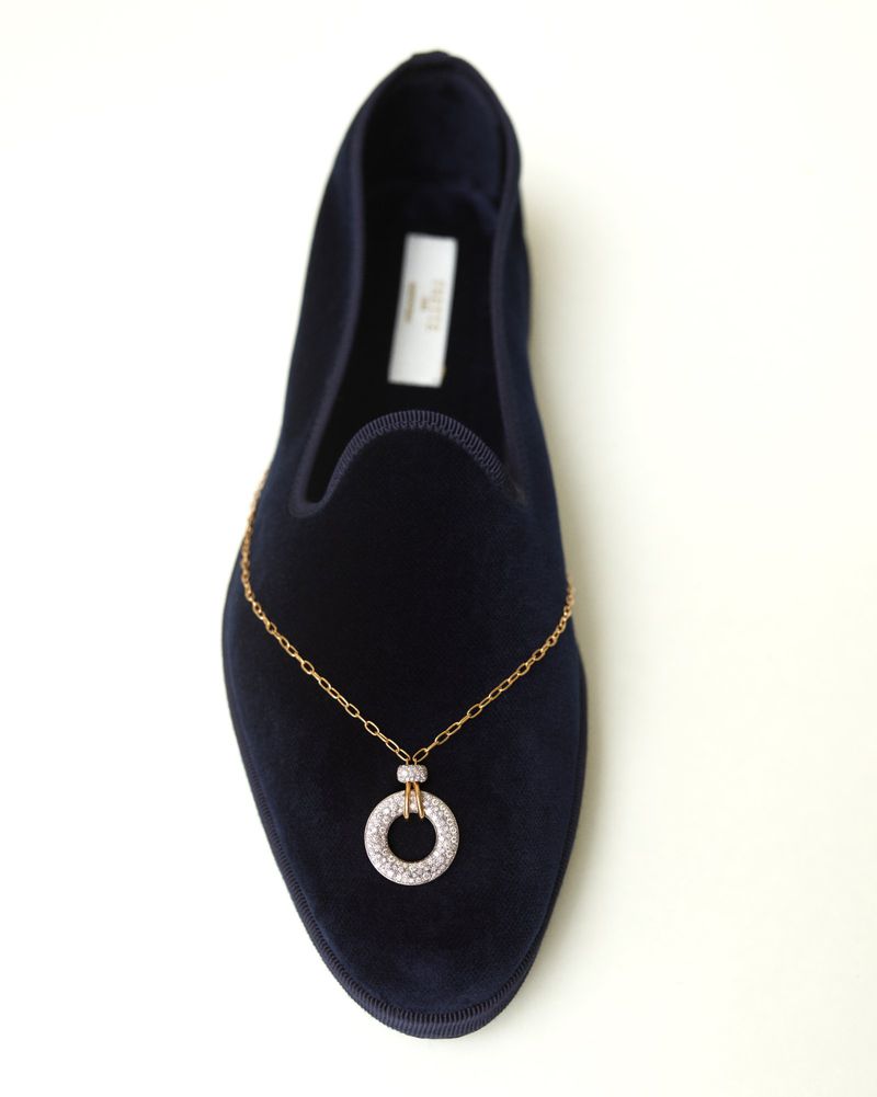 Close-up of a dark velvet loafer adorned with a delicate gold chain and a round diamond pendant.