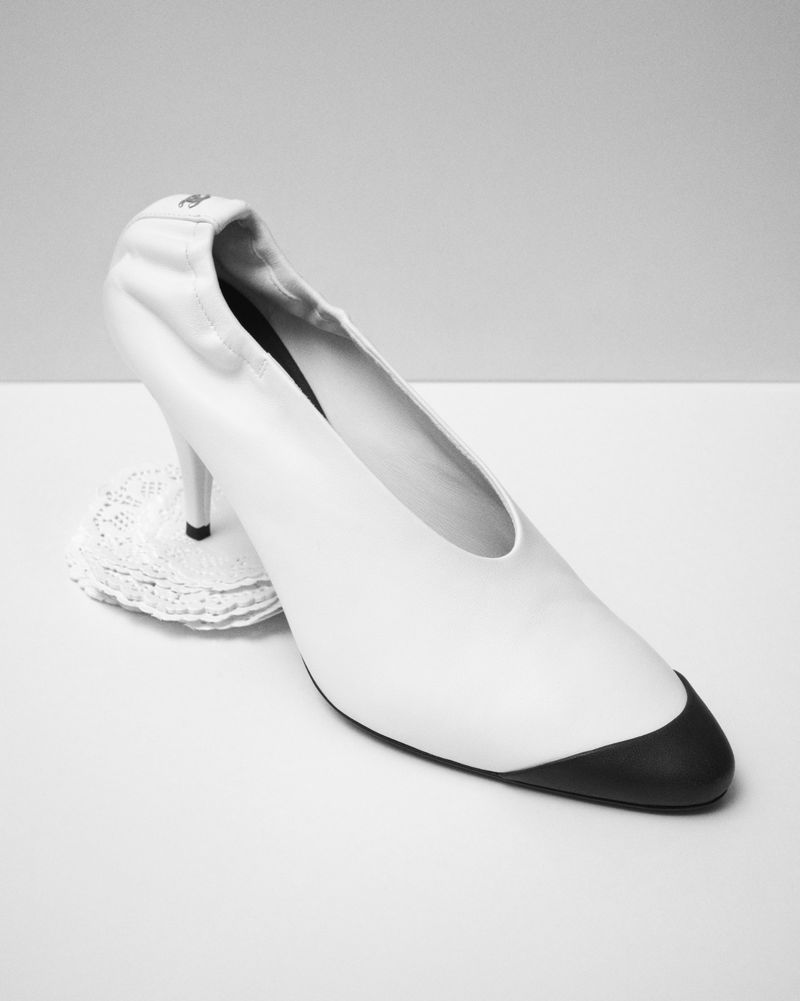 White pointed toe high heel shoe with black toe cap resting on a small white lace doily against a neutral backdrop