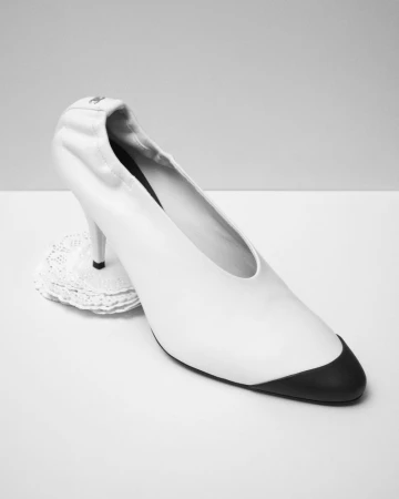 White pointed toe high heel shoe with black toe cap resting on a small white lace doily against a neutral backdrop