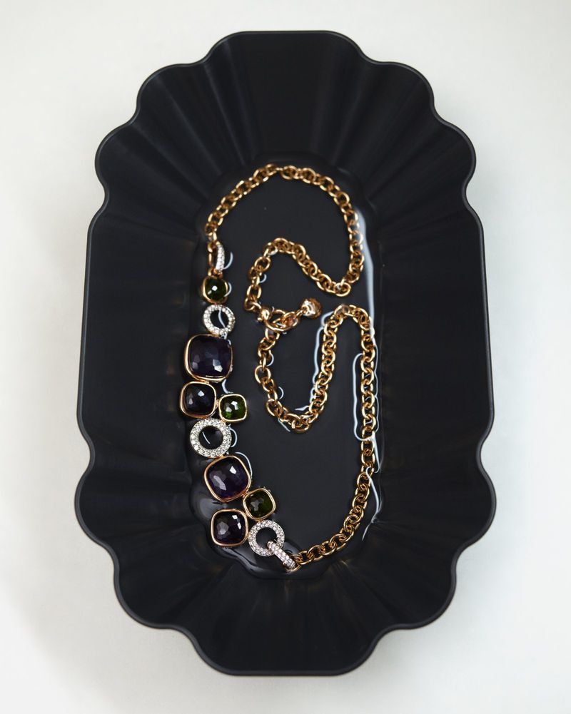 Gold statement necklace with dark purple and green stones resting on a black fluted tray.