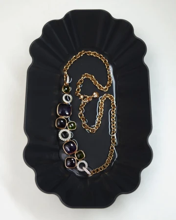 Gold statement necklace with dark purple and green stones resting on a black fluted tray.