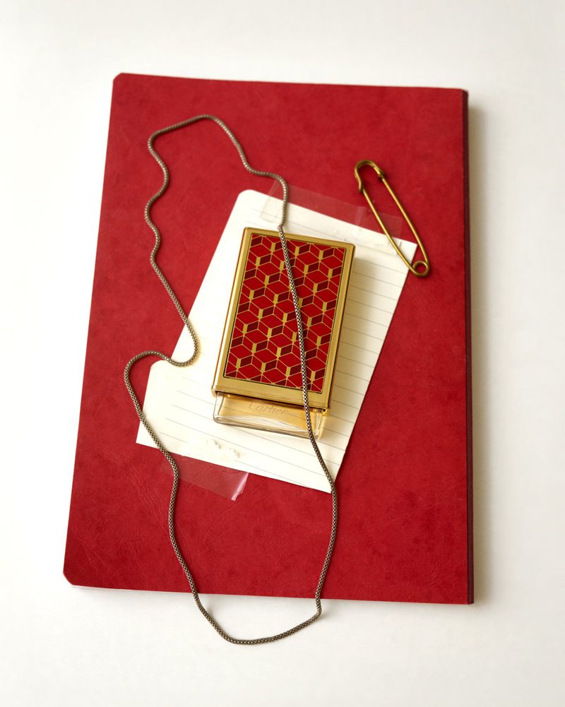 Overhead still life of a red journal, lined note card, gold geometric case, silver chain, and gold safety pin on white
