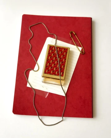 Overhead still life of a red journal, lined note card, gold geometric case, silver chain, and gold safety pin on white