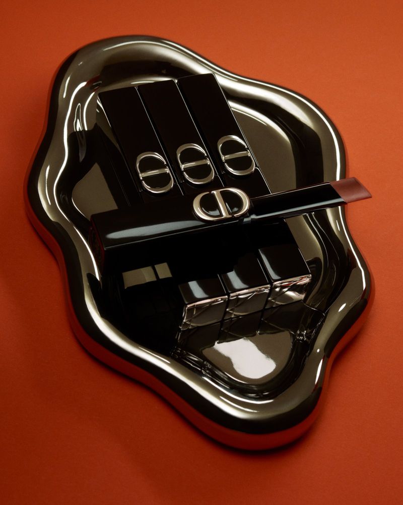 Several black lipstick cases arranged on a reflective wavy silver tray against a warm solid orange background.