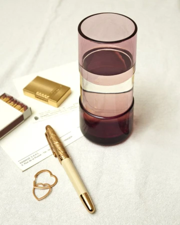 Cylindrical purple glass tumbler filled with water on a textured white fabric surface beside a gold pen and lighter.