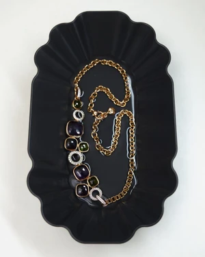 Gold statement necklace with dark purple and green stones resting on a black fluted tray.