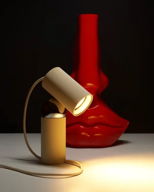 Modern beige desk lamp glowing beside a glossy red sculptural lip vase against a dark background
