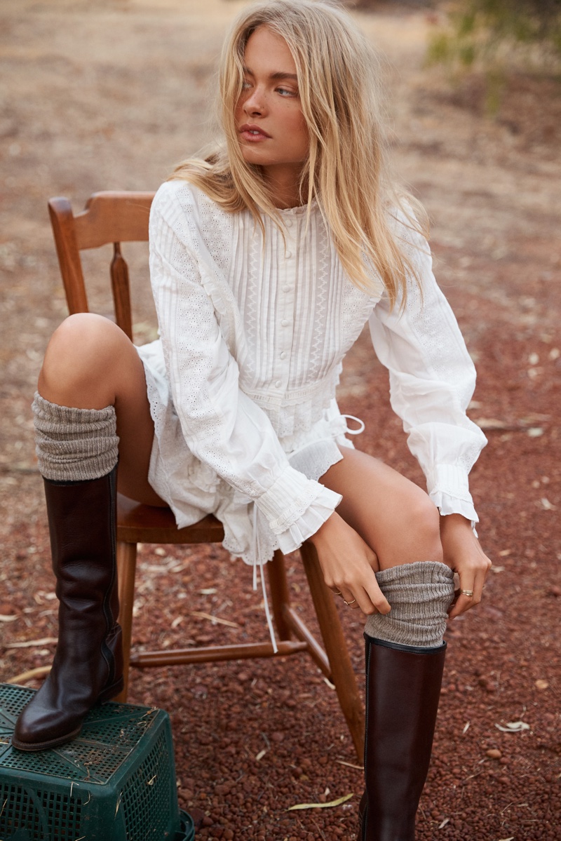 A blonde woman in a white dress and brown boots sits on a wooden chair outdoors with soft, diffused lighting.