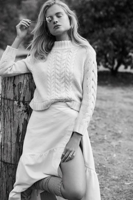 Black and white portrait of a woman holding a small flower to her eye, leaning against a wooden post outdoors.