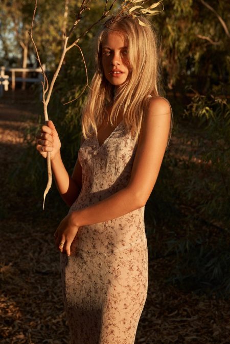 A fair-skinned woman poses in a floral dress outside, holding a slender branch, with greenery and a distant white fence in the background.
