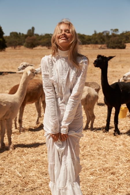 Woman in a white dress smiles in a field with a herd of alpacas, featuring a variety of colors in their coats and wool.