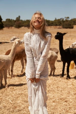 Woman in a white dress smiles in a field with a herd of alpacas, featuring a variety of colors in their coats and wool.
