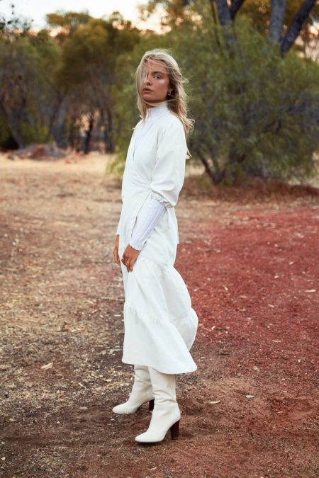 Full-length shot of an adult female in a long white dress and white boots, standing in a rural setting with sparse trees.
