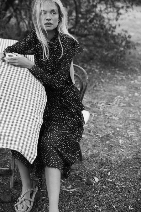 Black-and-white portrait of a fair-skinned woman in a polka dot dress sitting at a checkered table outdoors with foliage in background.