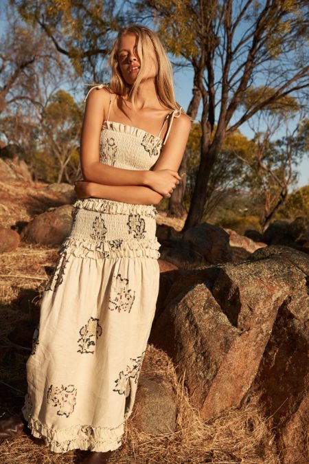 A woman poses in a smocked floral dress outdoors in the afternoon sun, framed by trees and reddish rocks, creating a natural backdrop.