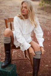 Blonde model sitting on a wooden chair outdoors wearing a white blouse and dark boots with grey knit leg warmers