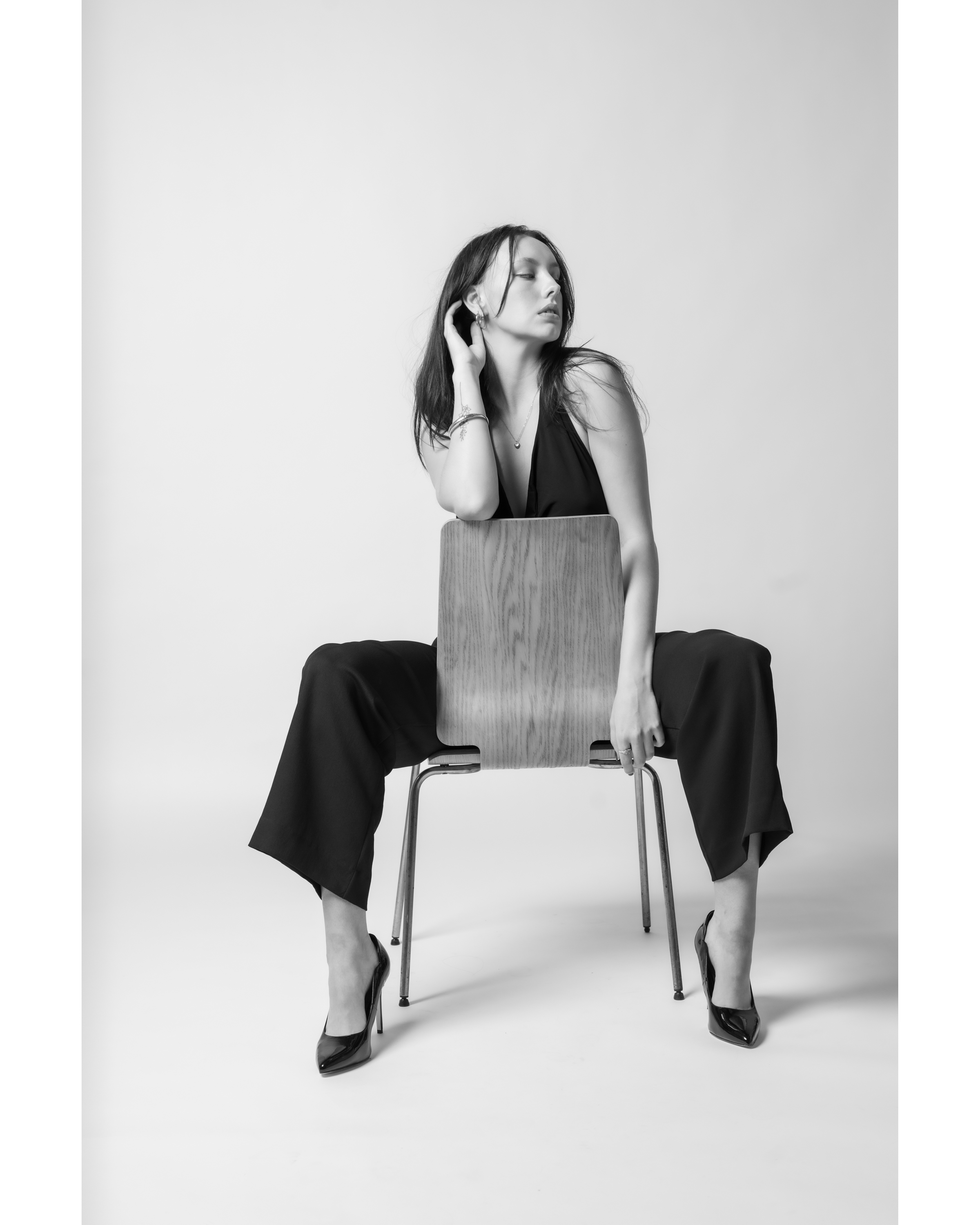 Black and white portrait of a woman in a dark halter jumpsuit seated backwards on a chair wearing heels in a studio setting.