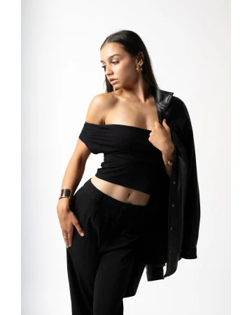 Portrait of a young woman in a black off-the-shoulder top and black pants, posing with a black jacket draped over her shoulder in a studio.