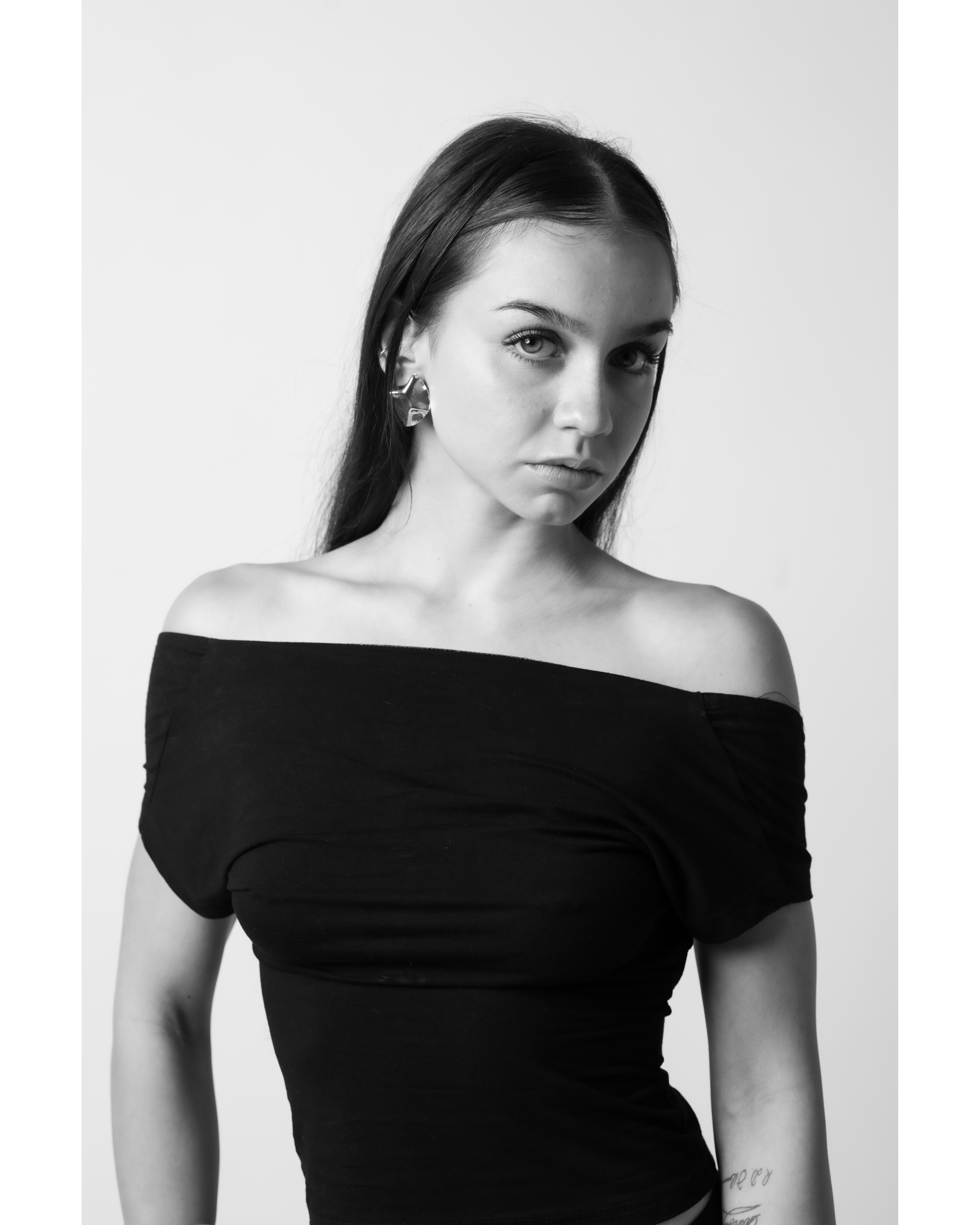 Monochrome portrait of a young adult woman with dark hair, wearing a black off-the-shoulder top, against a plain white backdrop in a studio setting.