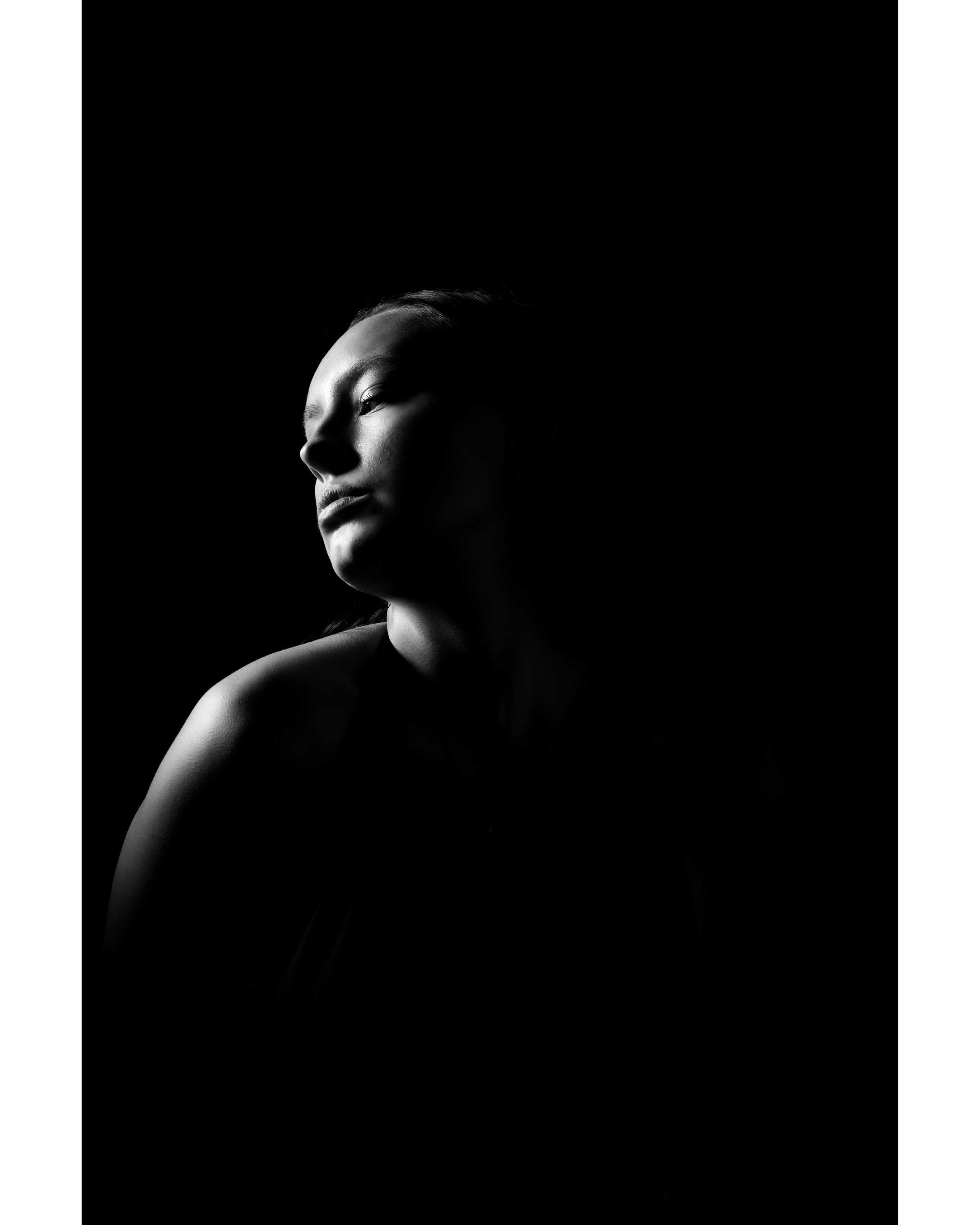 Monochrome portrait of a woman with soft lighting on her face against a stark black background, creating a dramatic chiaroscuro effect.