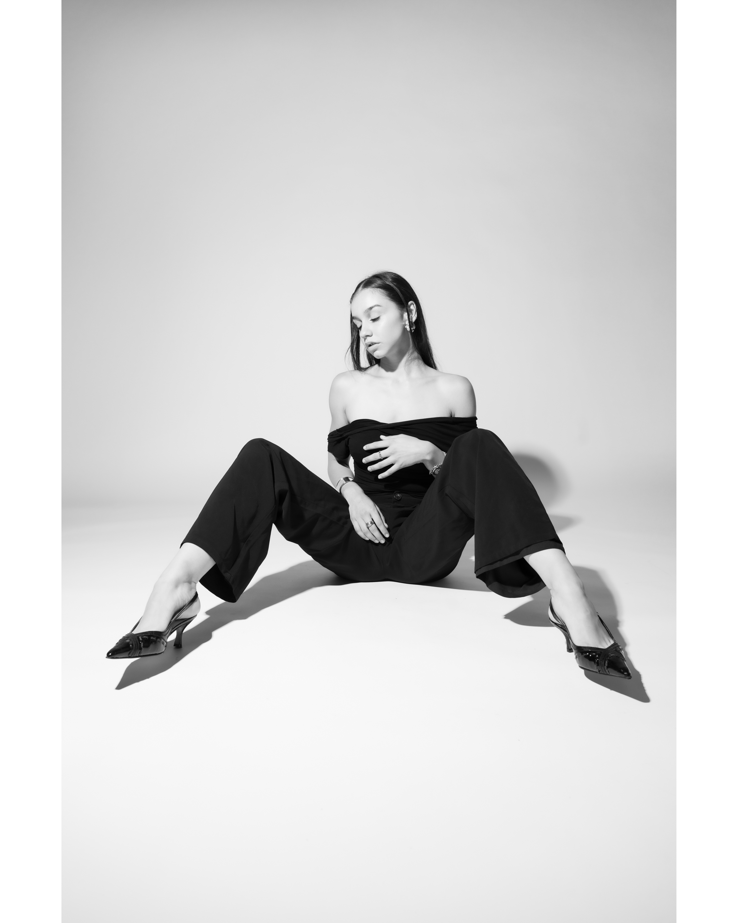 Monochrome portrait of a young adult woman in a black off-the-shoulder top and wide-leg pants sitting on a white surface wearing heels.