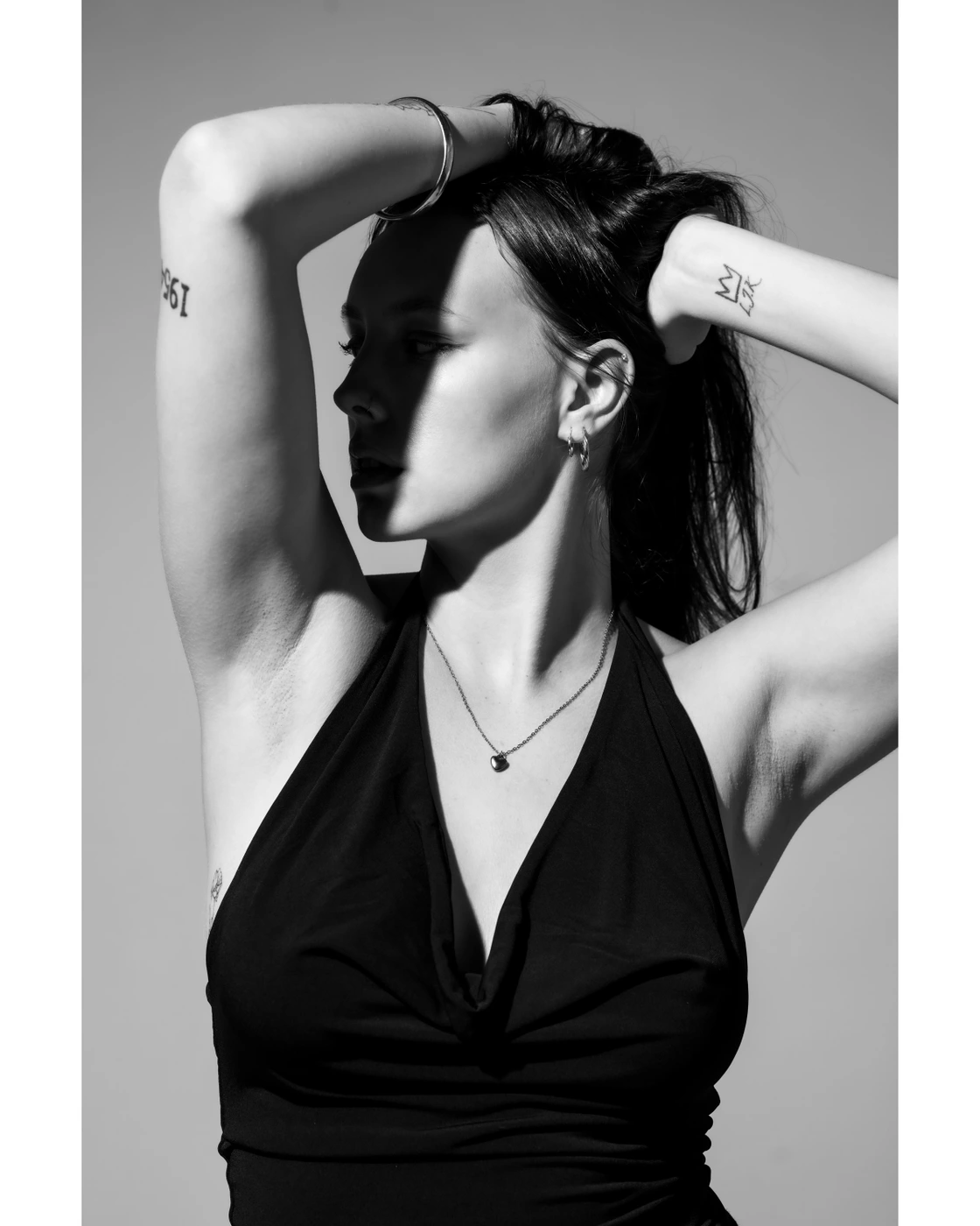 Black and white portrait of a model holding her hair up with both hands wearing a dark halter top.