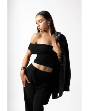 Model in black off-the-shoulder top and trousers holding leather jacket against a plain white background