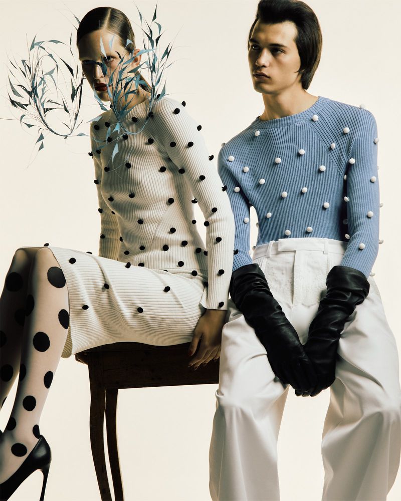 Fashion shot of a woman in a white dotted dress and a man in a blue sweater, white pants, and black gloves, posing against a light backdrop.