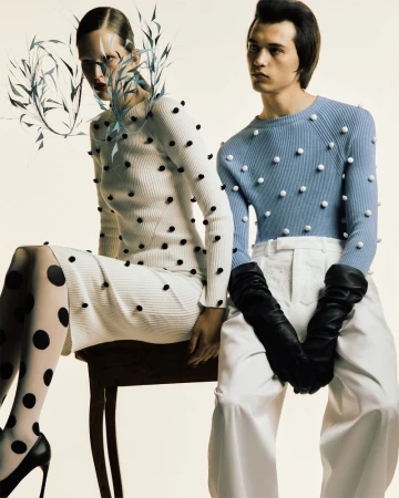 Fashion shot of a woman in a white dotted dress and a man in a blue sweater, white pants, and black gloves, posing against a light backdrop.
