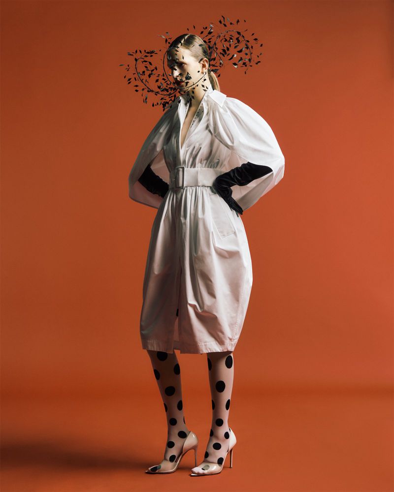 Fashion photo of a woman in a white dress with black polka dot stockings and heels, posing against a solid orange backdrop.
