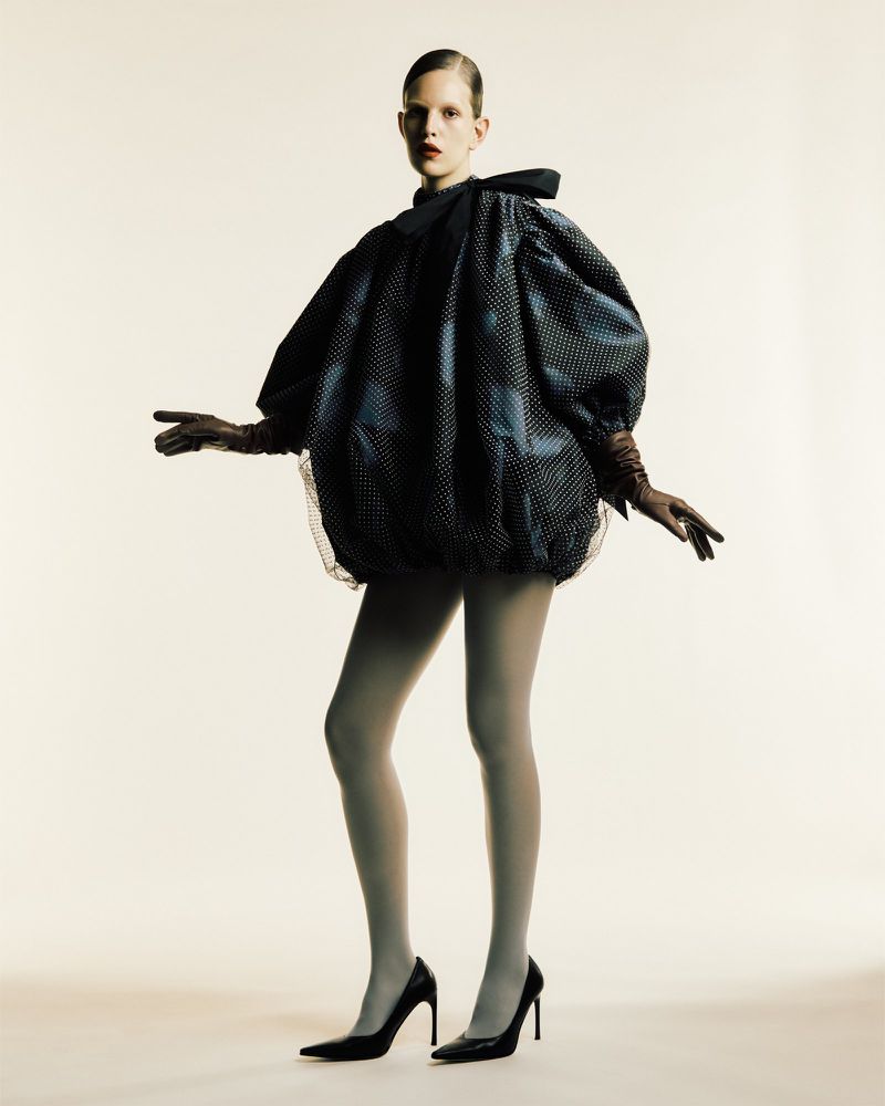 Fashion photo featuring a female model in a polka dot dress, gloves, tights, and heels, posing against a neutral backdrop with soft studio lighting.