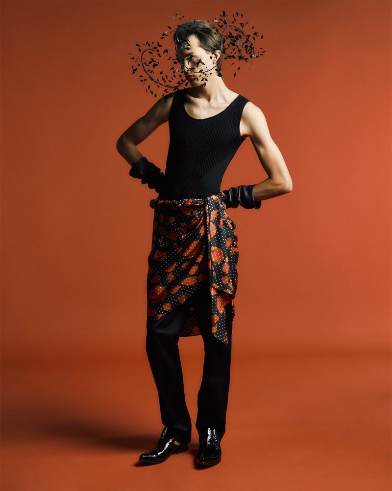 A slender man in a black top, patterned wrap, black pants, and gloves poses against an orange backdrop, adorned with decorative black leaves.