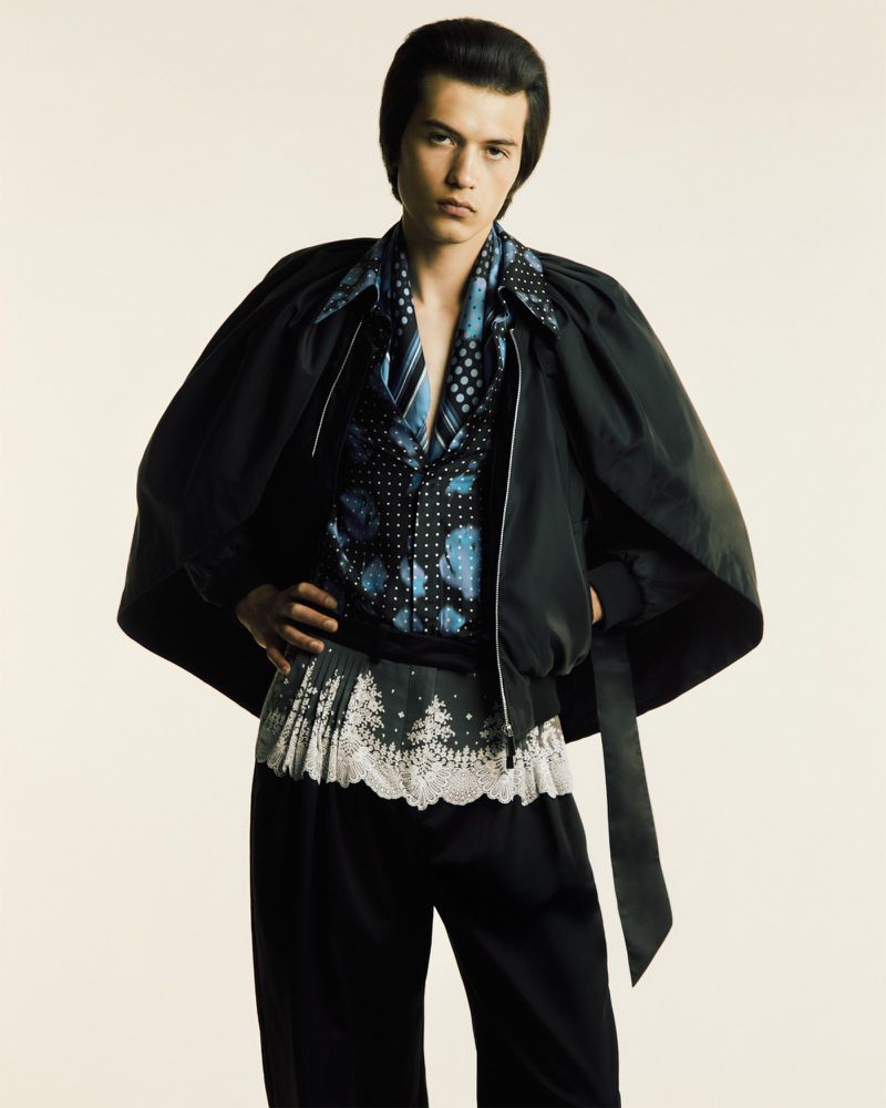 Portrait of a person posing in a black jacket, patterned top, and black trousers against a neutral backdrop in a studio setting.
