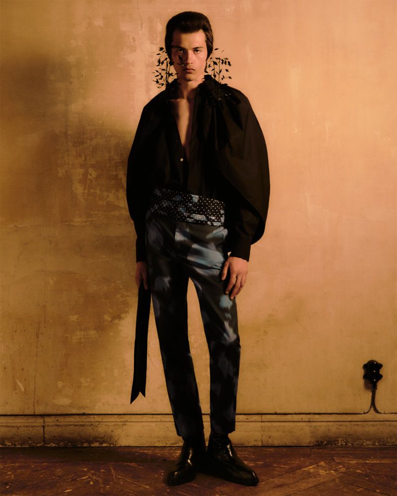 A young adult male poses in a studio wearing a black shirt with oversized sleeves, patterned pants, and shiny black shoes, against a textured wall.
