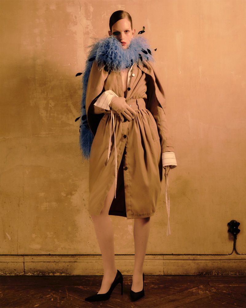 Fashionable woman in tan coat and blue feather boa poses in studio. Black heels, gloves, and dramatic lighting complete the editorial look.
