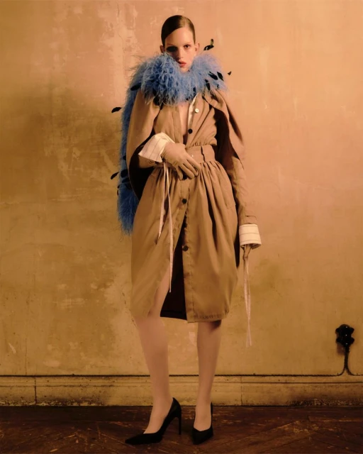 Fashionable woman in tan coat and blue feather boa poses in studio. Black heels, gloves, and dramatic lighting complete the editorial look.