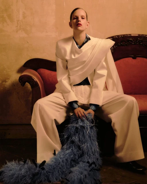 Woman in white pantsuit sits on red couch holding feather boa in studio with warm lighting. Fashion editorial portrait.