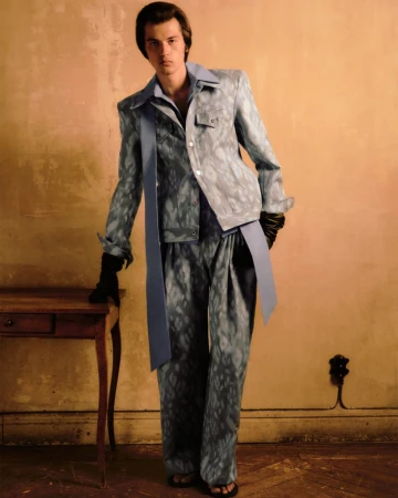 Full-length portrait of a young adult male fashion model in a patterned blue suit with a sash and gloves, posing in an indoor setting.