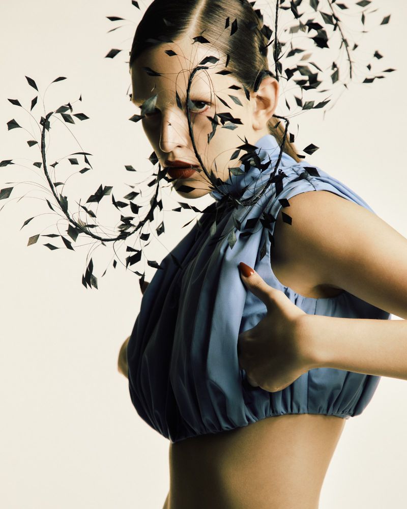 Fashion editorial shot of a woman wearing a blue top with a black, leafy headpiece, posing in front of an off-white background in studio lighting.