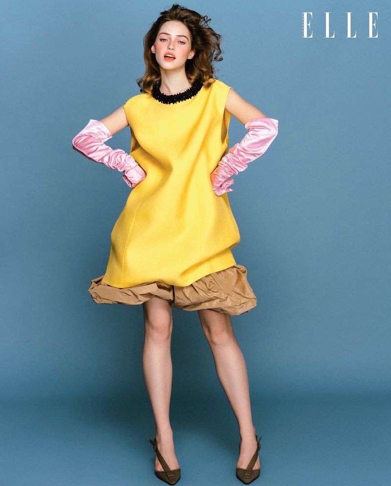 Model in a bright yellow dress with pink satin gloves and brown trim against a blue backdrop for an Elle Singapore editorial.