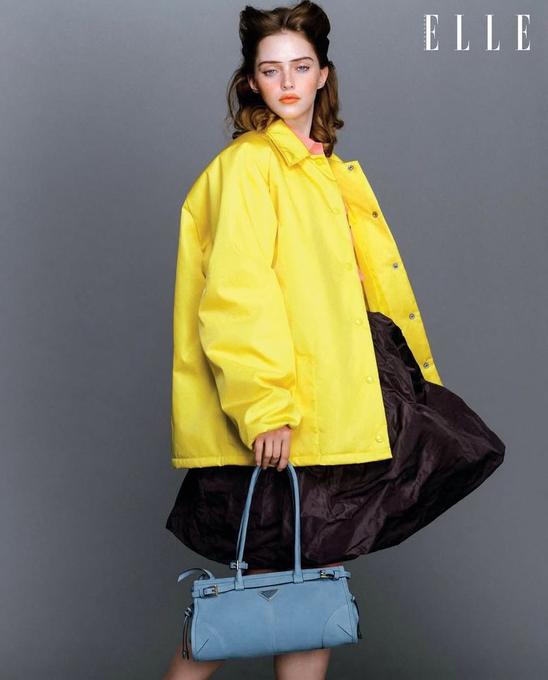 Model wearing a bright yellow jacket and a dark skirt holding a light blue handbag against a grey backdrop