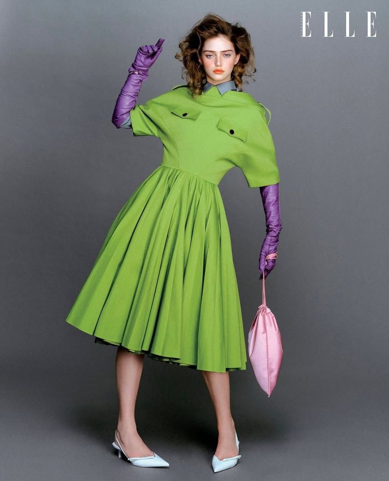 Model in green dress with purple gloves and pink handbag against a grey backdrop for Elle Singapore fashion editorial