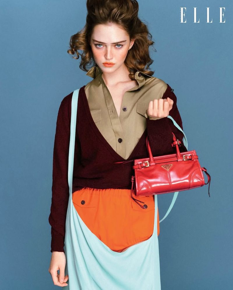 Model wearing a layered brown top and light blue skirt holding a red handbag against a blue background for Elle Singapore.