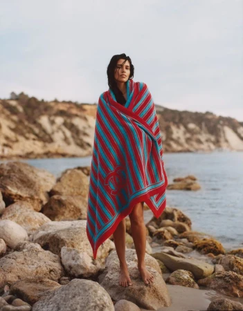 Model Pooja Mor wrapped in a striped red and blue blanket standing on a rocky beach for Grazia UK