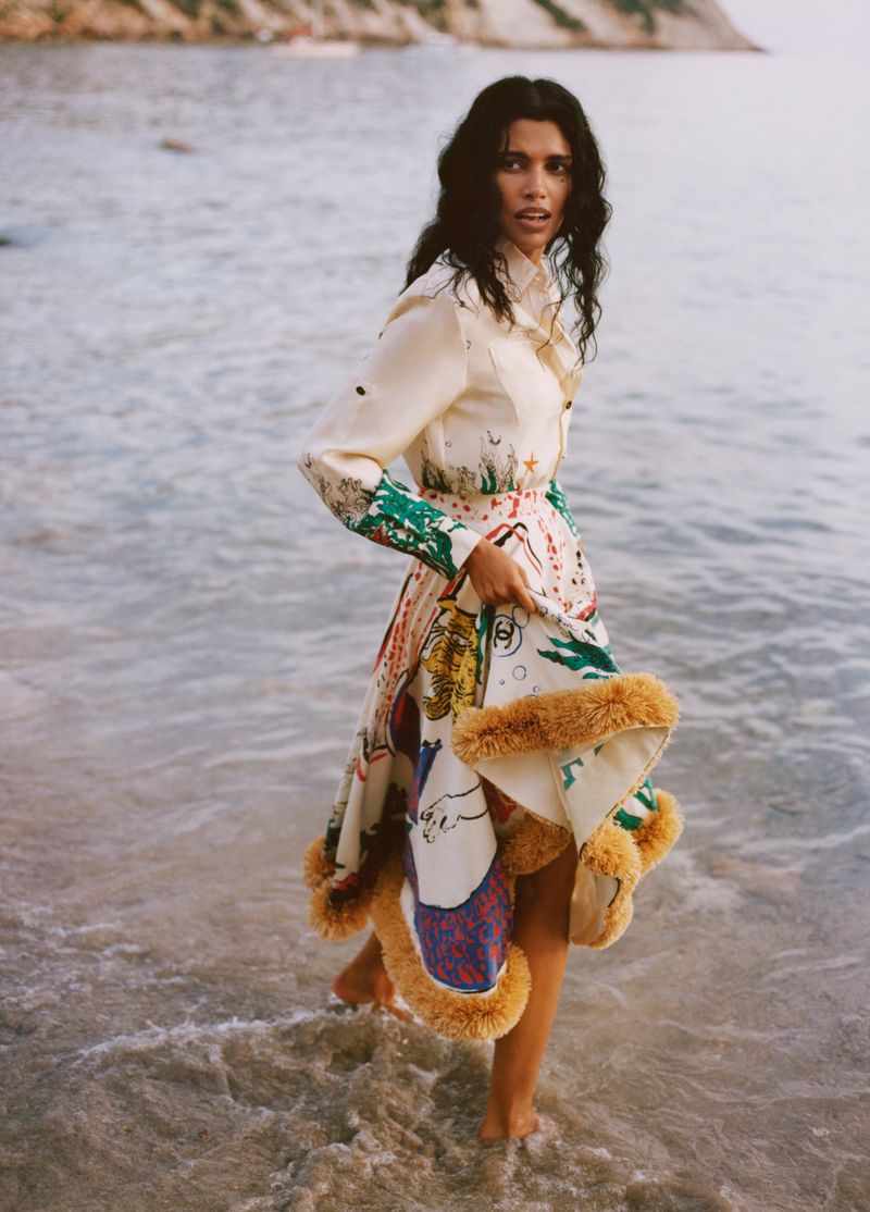 Model Pooja Mor in a printed dress with fur trim walking in shallow ocean water for Grazia UK Shore Things editorial