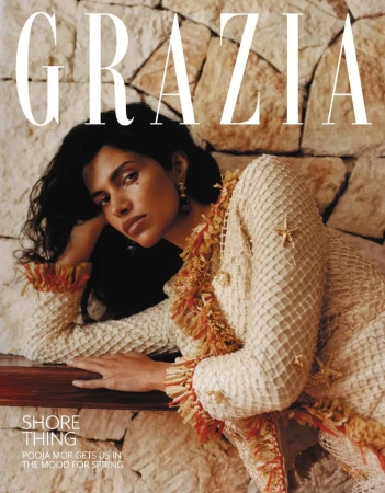 Model Pooja Mor leaning against a rustic stone wall wearing a textured cream dress for Grazia UK magazine.